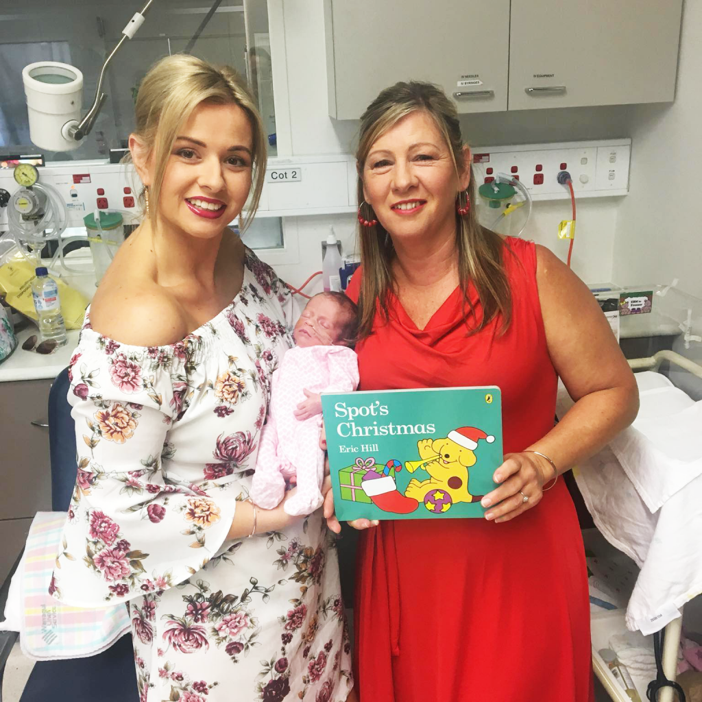 Young mum in wearing floral dress in a hospital ward holding her tiny premature baby next to a woman holding the book 'Spot's First Christmas'. 